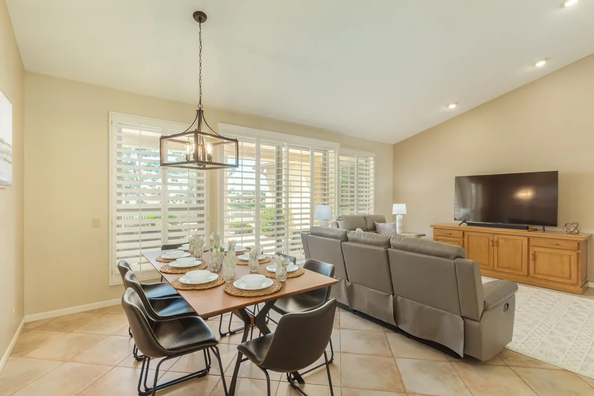 This interior shot features a spacious open-concept living and dining area with a reclining sofa, a large television unit, and a dining table set for a meal. Large windows with shutters allow abundant natural light to fill the space, and the overall mood is inviting and comfortable, perfect for gatherings and relaxation.