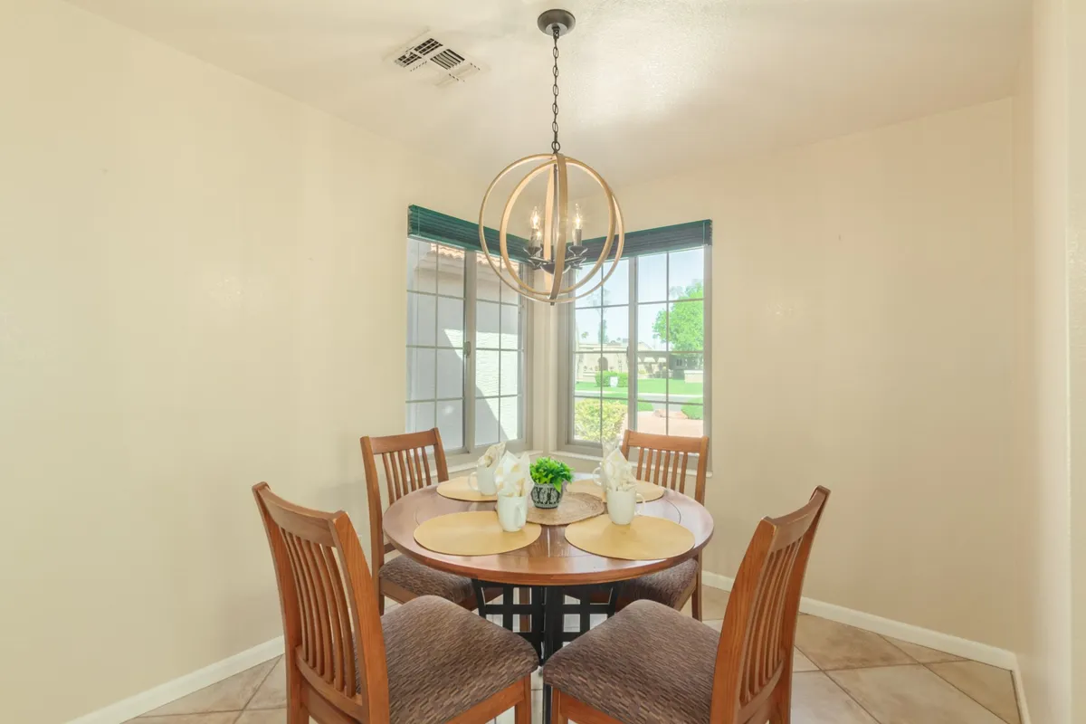 A cozy breakfast nook featuring a round dining table beneath a sparkling chandelier, surrounded by windows that let in natural light and offer a view of the outdoors. A decorative plant adds a welcoming touch.