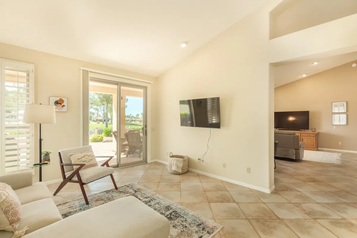 A comfortable living room scene with a sofa, television, armchair, area rug, and a prominent sliding glass door that opens to the outdoors. The design is relaxed yet stylish, and the glass door frames views of the green surroundings, emphasizing the indoor-outdoor connection.