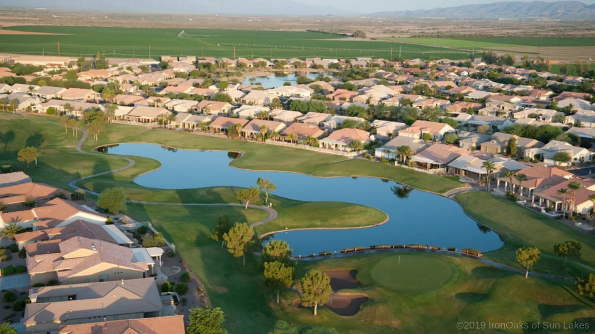 A panoramic view of a lush golf course bordered by tranquil ponds, with well-kept lawns and residential homes lining the perimeter. The landscape is bathed in gentle sunlight, evoking a sense of relaxation and community.