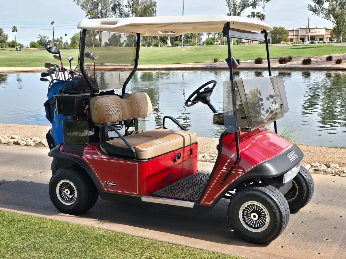 A golf cart is parked on the green grass near a water body and golf course, with golf clubs visible and open skies above. The scene is cheerful and active, hinting at leisurely rounds of golf, convenient access, and the vibrant country club lifestyle.