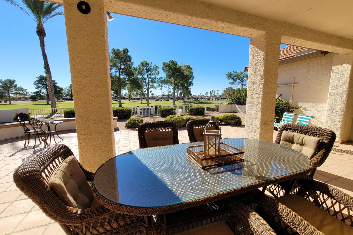 A beautiful rattan dining table set for eight sits on a broad patio, adorned with a lantern centerpiece. The space is surrounded by lush greenery and palm trees, with a sweeping view of the distant golf course. The late afternoon light glows warmly, evoking the perfect setting for sunset dinners or morning coffee.