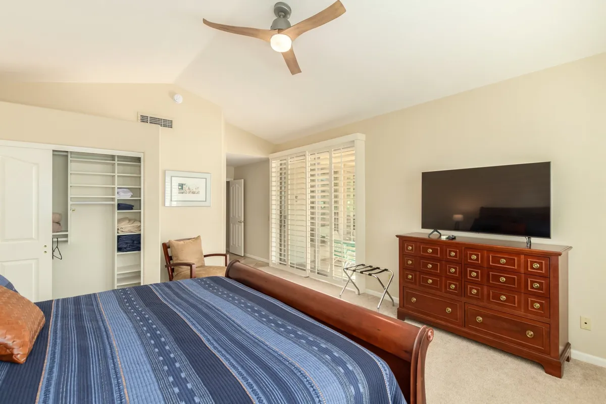 A serene bedroom featuring a neatly made bed, a dresser with a TV on top, a closet, and a comfortable chair. The room is accented by natural light filtering through the windows and calming, neutral decor.