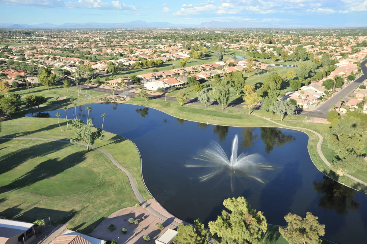 A tranquil lakeside setting featuring a sparkling fountain at the center, surrounded by lush greenery and glimpses of the community's well-maintained residential areas. The walking paths offer scenic views, with residents picnicking by the lake or enjoying morning walks along the peaceful shoreline.